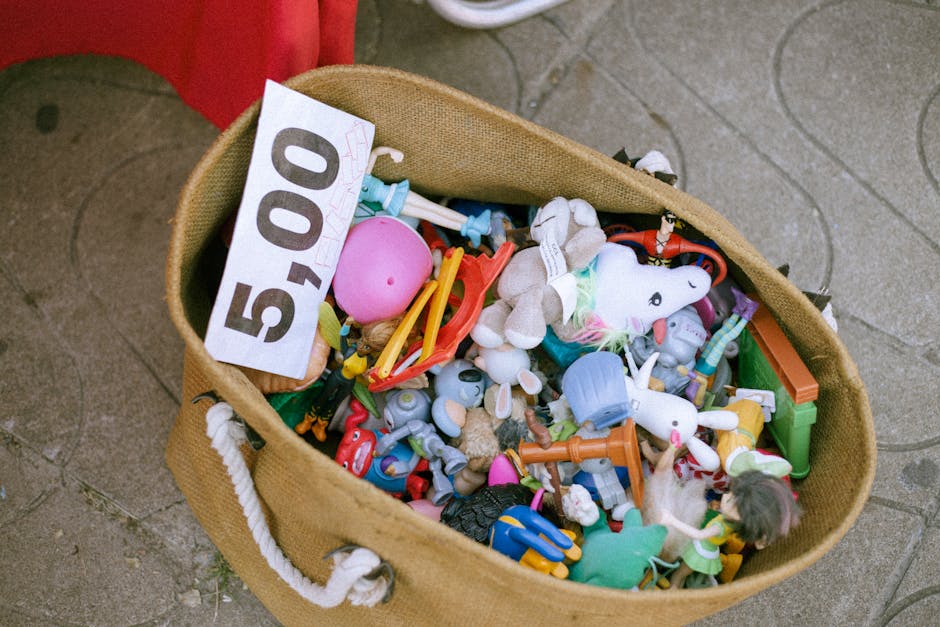 A tan, woven fabric tote bag with white rope handles is placed on a paved surface, filled with an assortment of children's toys and plastic items. Visible objects include a pink rubber ball, a white unicorn plush with a rainbow-colored mane, and various small plastic figures and figures, some with brightly coloured clothing and accessories. The toys are made from smooth plastic and fabric, displaying textures ranging from shiny and glossy to soft and fuzzy. The bag leans slightly to one side, revealing the different shapes and sizes of the toys, which are loosely packed inside. The paved surface underneath features rounded stone patterns, suggesting an outdoor environment such as a market or street scene. The overall scene appears to be part of a rubbish clearance or alternative waste handling scenario, with Waste Removal Tooting providing an efficient way to collect and remove such miscellaneous waste. The image emphasizes the importance of private disposal services for non-standard waste items like children's toys and clutter collected for rubbish removal.