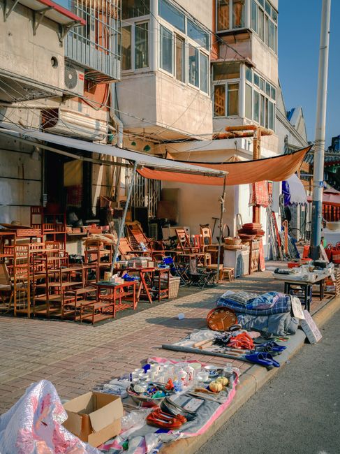 The image depicts a street scene outside a building with large glass windows and a concrete facade, likely in an urban area. In the foreground, various second-hand items are laid out on the pavement, including plastic bottles, small household goods, and miscellaneous objects arranged on and around a table covered with a cloth, suggesting a street market or clearance sale. To the right, a pile of larger items such as a rolled-up mat, a cardboard box, and other discarded household objects can be seen, indicating ongoing rubbish clearance or removal activities. Behind the display, outdoor tables and wooden chairs, some stacked and others arranged singly, are visible under a partially extended fabric awning, providing shade for the market stall. The building has a mix of metal railings, pipes, and air conditioning units attached to the exterior, with multiple balconies overhead. The scene is illuminated by natural daylight, capturing the textures of the mix of household items, the urban environment, and the activities associated with private waste collection or rubbish clearance services, exemplified by Waste Removal Tooting.