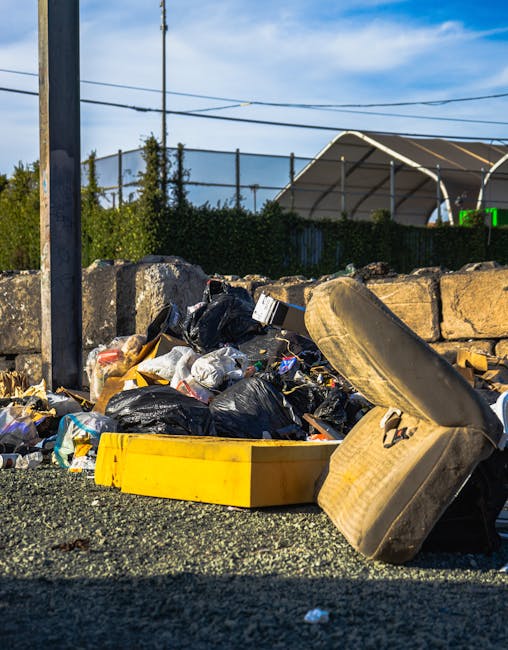 A close-up view of unmanaged rubbish piled on a gravel surface near a stone wall, including black plastic garbage bags, a yellow plastic container, a worn beige car tyre leaning against the bags, and various miscellaneous waste items. In the background, there is a metal fence, green foliage, utility wires, and a partly cloudy blue sky, suggesting an outdoor site possibly used for private waste disposal or clearance by Waste Removal Tooting. The scene highlights an area where non-recycled waste materials, typical of a rubbish removal service, have been accumulated temporarily, emphasizing the need for professional clearance or waste management solutions. The natural lighting reveals textures of the waste, including the glossy surface of plastic bags and the rough rubber of the tyre, under a bright daylight setting. This image exemplifies an instance where independent rubbish collection or on-site clearance might be required, aligning with waste removal activities in a residential or commercial context.