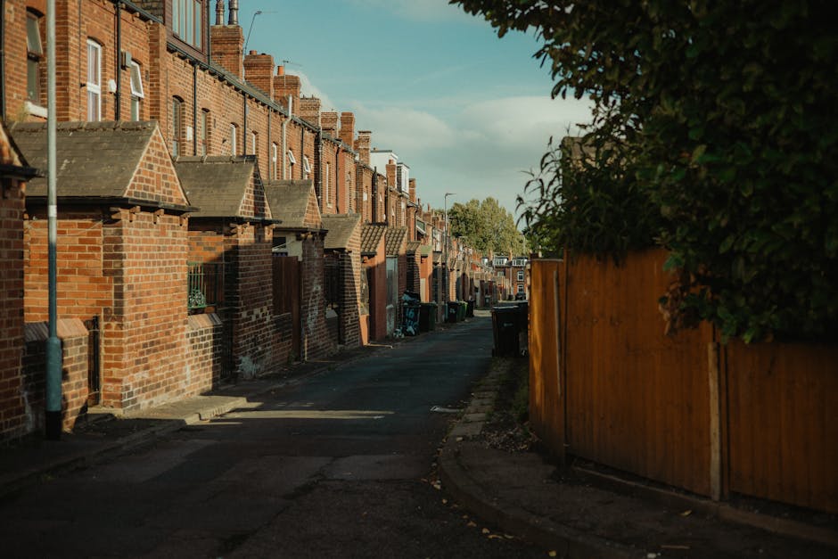 A quiet residential alleyway lined with traditional red-brick terraced houses on the left, each with small front yards enclosed by low brick walls and sloped tiled roofs, illuminated by warm late-afternoon sunlight. To the right, a tall, natural green hedge partially obscures a wooden fence that runs along the side of the alley, separating the private gardens from the walkway. The narrow paved pathway curves gently and is bordered by a raised concrete curb on one side and a strip of pavement on the other. Several black wheelie bins are positioned near the doorways of the houses, indicating bin collection points typically used for waste disposal. The scene is clean and orderly, with a clear blue sky and some scattered clouds, capturing a typical urban environment that may require services such as waste removal or private rubbish collection efforts by companies like Waste Removal Tooting. The photograph’s lighting and shadowing suggest late afternoon, creating a calm atmosphere characteristic of a well-maintained residential area, possibly involving private waste management rather than council bin collections.