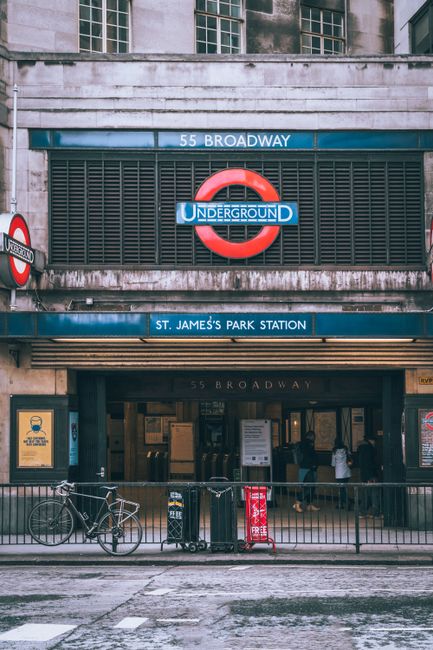 The exterior entrance of St. James's Park Station, an underground railway station located at 55 Broadway, featuring a prominent London Underground roundel sign with a red circle and blue bar displaying the station name. Above the entrance, there are metal louvers and concrete detailing, with the address '55 BROADWAY' inscribed at the top. The station signage includes blue panels with white text and a smaller blue sign indicating the station's name beneath the main sign. At street level, there is a sidewalk with a metal railing, a bicycle parked on the left, and a red and black trash bin positioned near the railing. Partially visible behind the railing are pedestrians entering the station. The scene is lit with natural daylight, capturing a typical urban environment associated with London public transportation infrastructure, relevant for discussions around public or alternative waste management services like rubbish removal or street cleaning near busy transit locations.