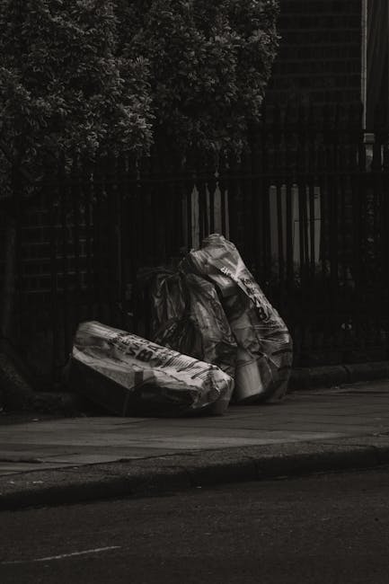Three large black plastic rubbish bags, resting against a dark metal fence on the edge of a paved street, with dense foliage in the background. The bags appear to be filled with household or garden waste, with visible creases and slight bulges indicating their contents. The bags are positioned on the sidewalk, partially overlapping, and are situated near a curb, suggesting a private collection or disposal scene. The environment appears subdued with minimal natural light, casting subtle shadows around the bags, consistent with an urban or suburban setting where waste removal services such as those offered by Waste Removal Tooting might handle such refuse. The scene emphasizes the importance of professional rubbish removal to manage waste efficiently and maintain area cleanliness, aligning with services for independent clearance outside conventional municipal collection.
